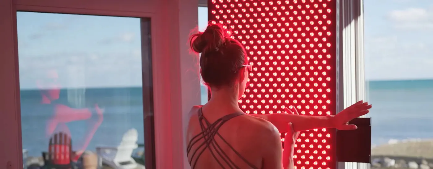 A modern home wellness setup showing a Hooga full-body red light therapy panel and a person using a handheld device on their face